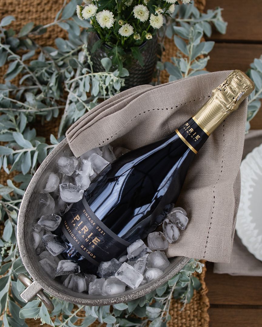 Bottle of Pirie sparkling wine chilling in a silver ice bucket with ice cubes, a linen napkin, and eucalyptus and white flowers nearby.