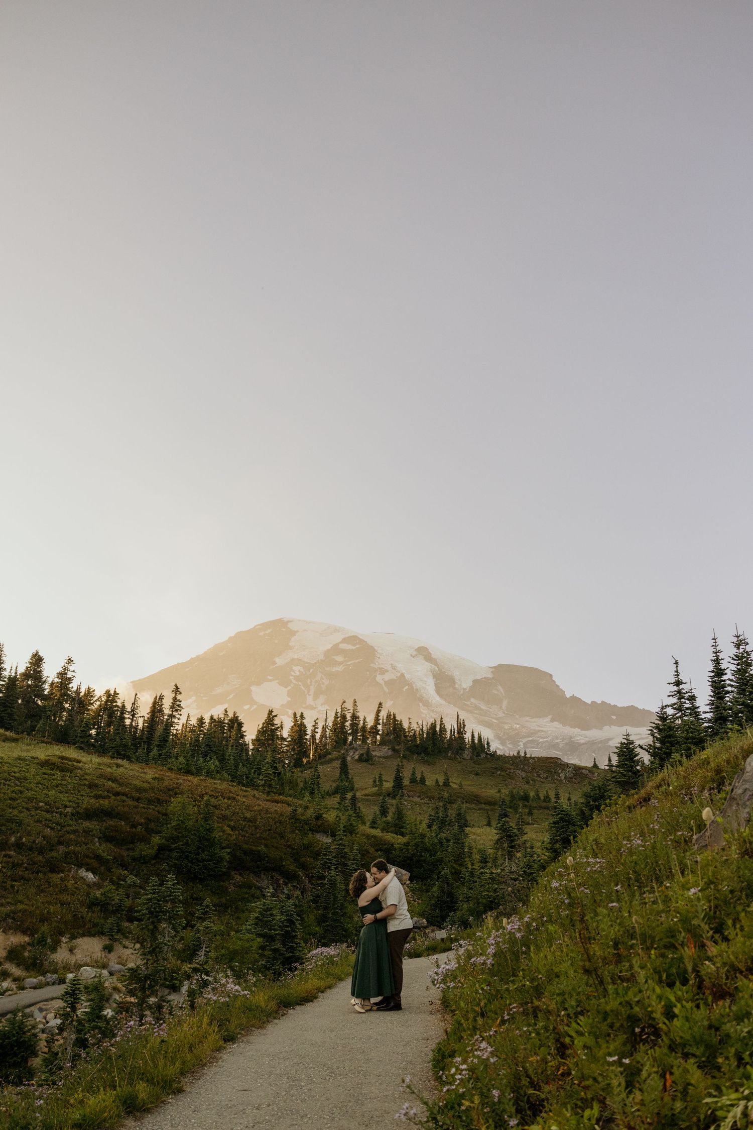 Couple embracing on a mountain trail at sunset with snow-capped Mount Rainier and evergreen trees in the background.