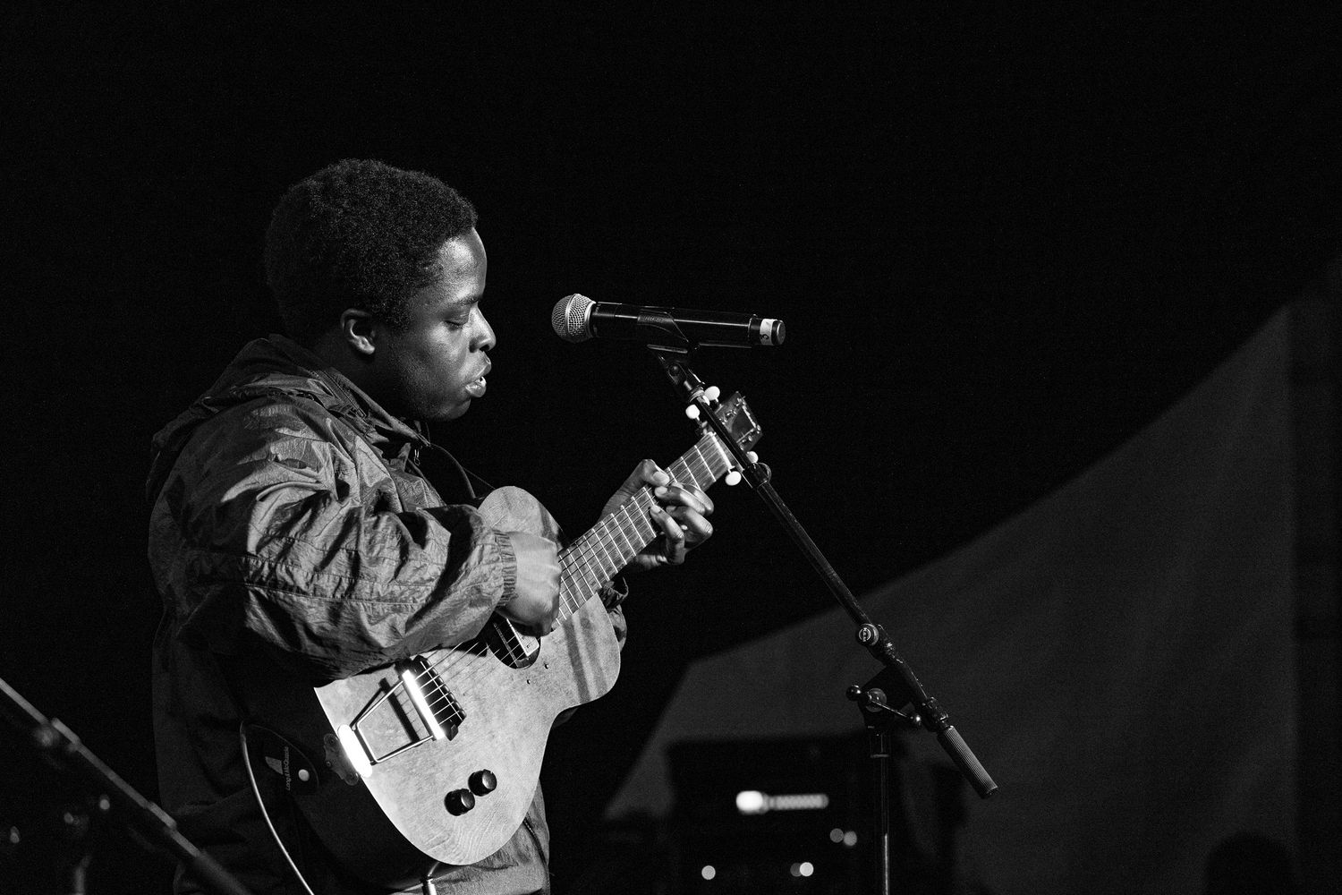 Black and white photo of a musician playing electric guitar and singing into a microphone on stage.