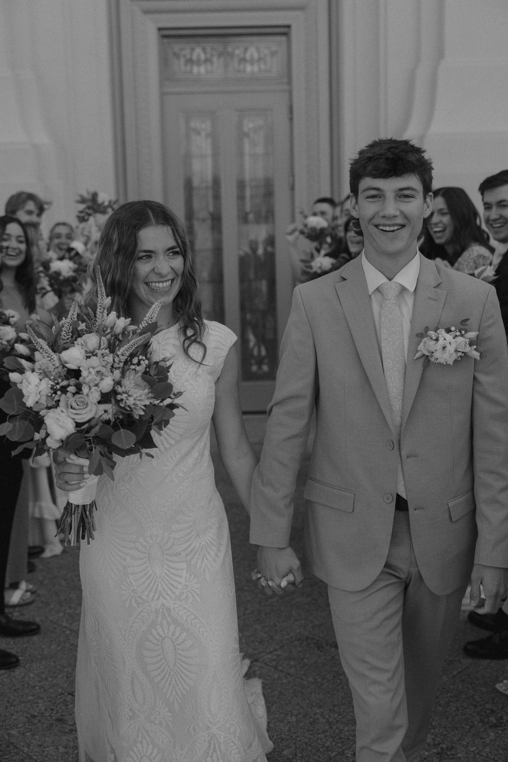 Black and white photo of smiling newlyweds exiting temple doors surrounded by cheering guests.