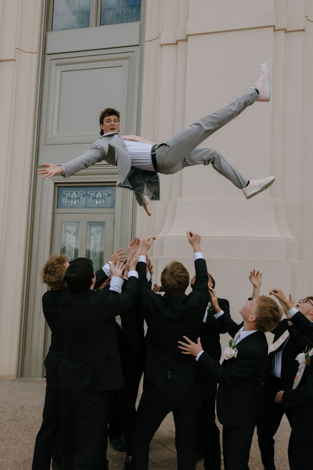 Groomsmen in black suits tossing groom in gray suit into the air outside a wedding venue.