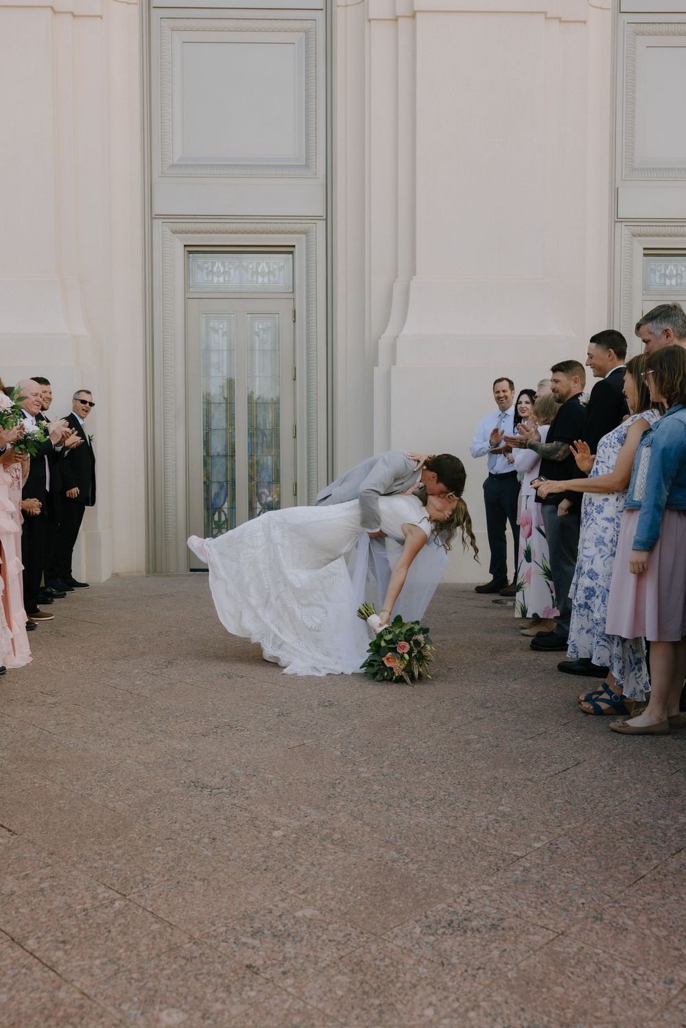 Groom dipping bride in dramatic kiss outside temple entrance while wedding guests look on.