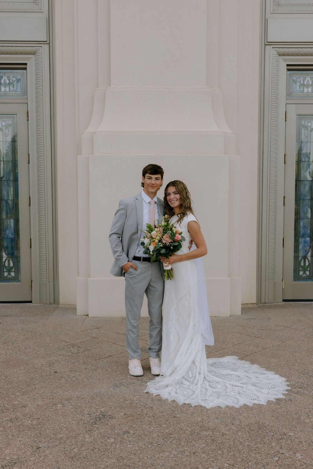 Bride and groom pose together in front of a white building, she holds a colorful bouquet in her lace wedding gown.