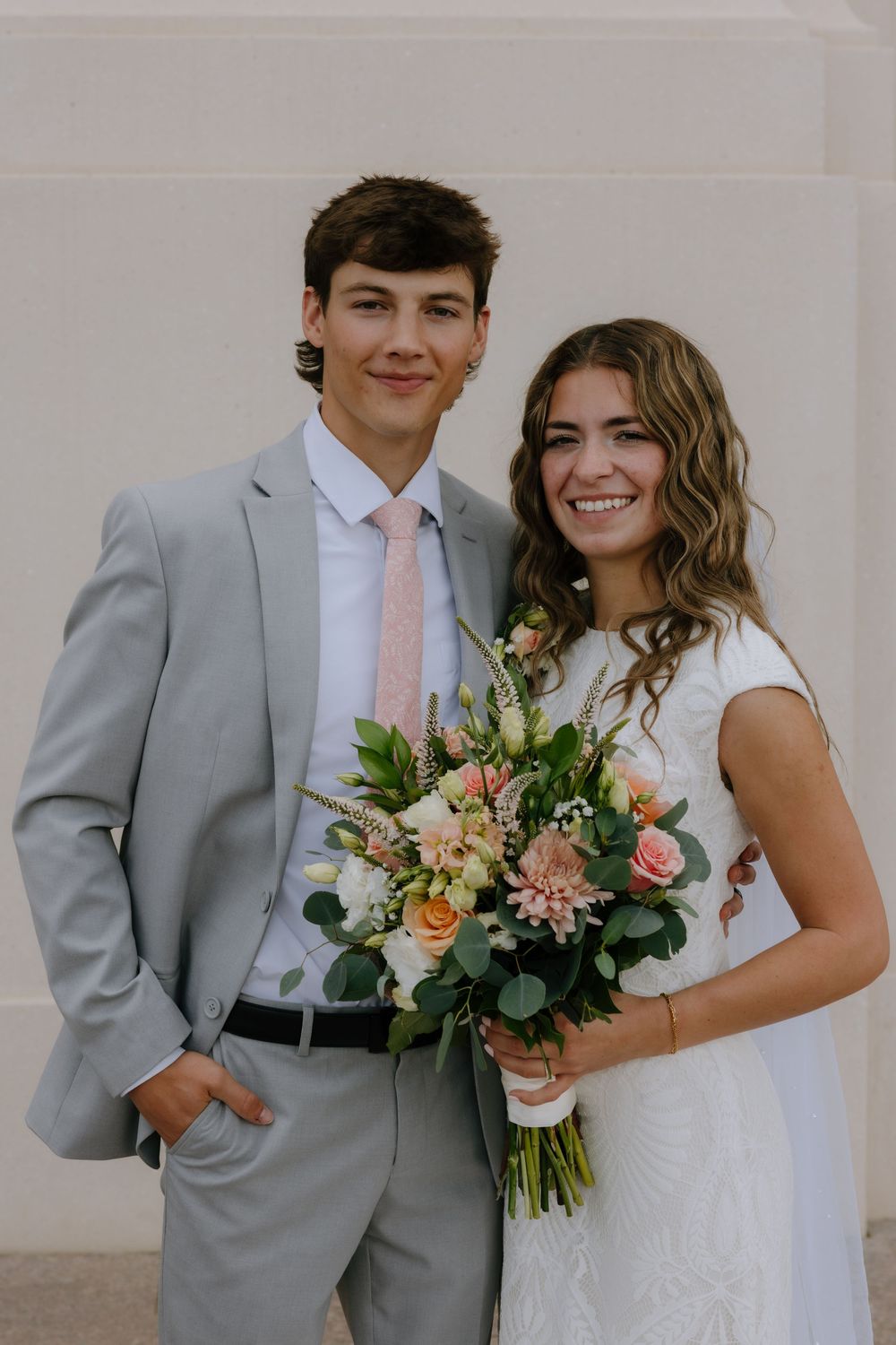 Smiling bride and groom pose together, she holding a peach and white bouquet, both in wedding attire outdoors.