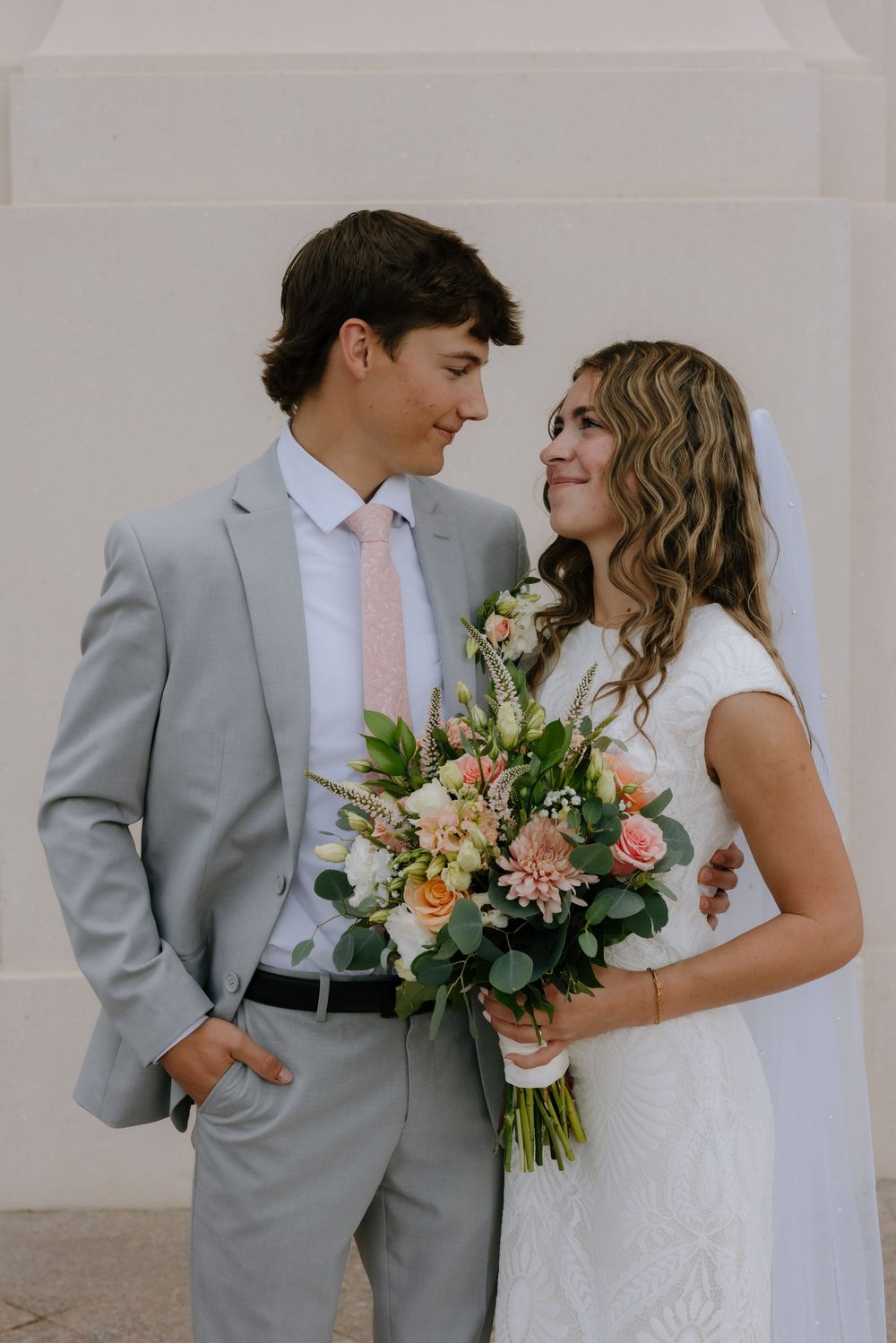 Bride and groom in gray suit gazing at each other while holding a peach and pink bridal bouquet.