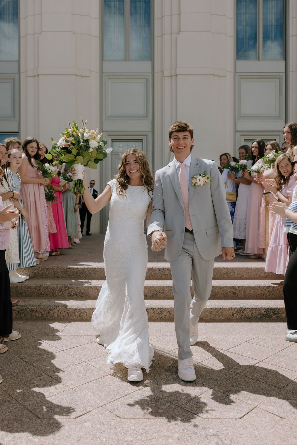 Joyful newlyweds descend temple steps holding hands, bride raising bouquet, guests lining the path cheering.
