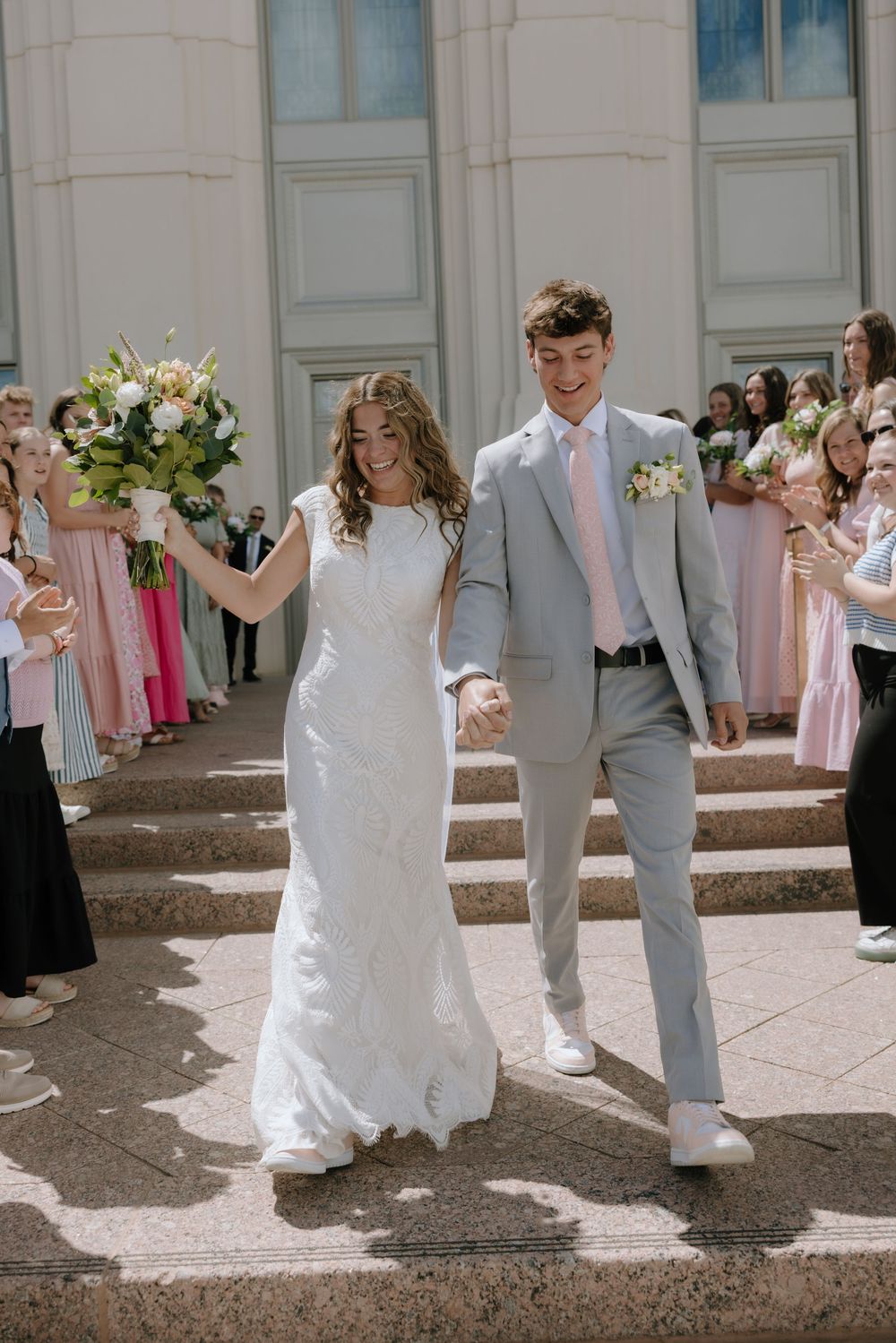 Joyful bride and groom descending temple steps hand-in-hand while guests celebrate around them.