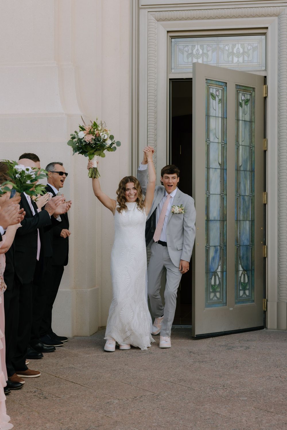 Joyful bride raising bouquet as couple exits LDS temple doors surrounded by applauding wedding guests.