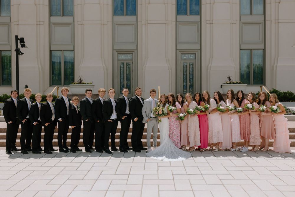 Wedding party posing outside a grand building, groomsmen in black tuxedos and bridesmaids in pink dresses flanking the bride and groom.