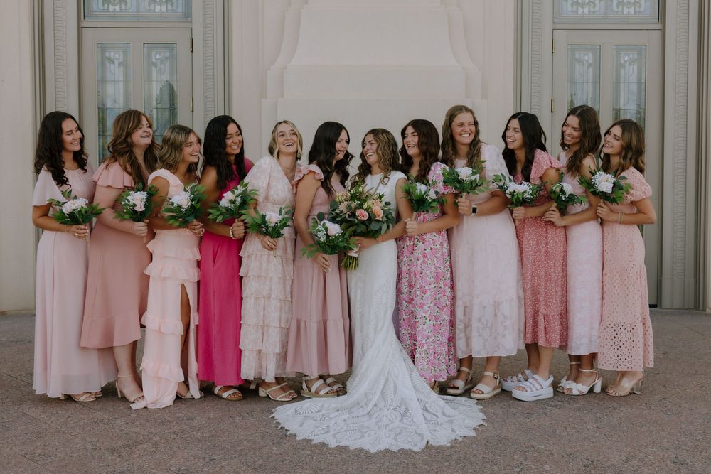 Bride in lace gown poses with bridesmaids in mismatched pink dresses holding bouquets outdoors.
