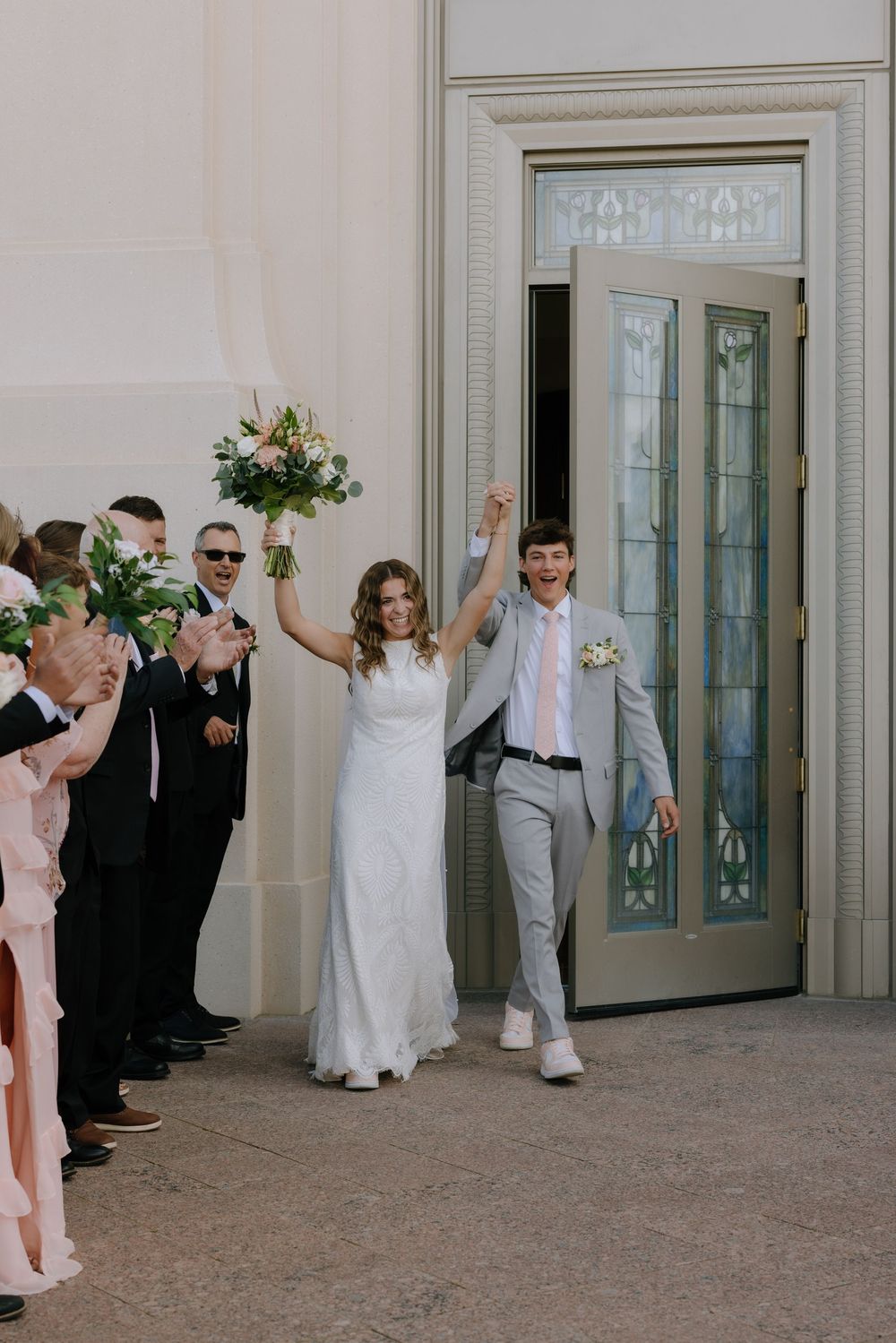Newlyweds celebrating exit from LDS temple doors with raised hands and bridal bouquet.