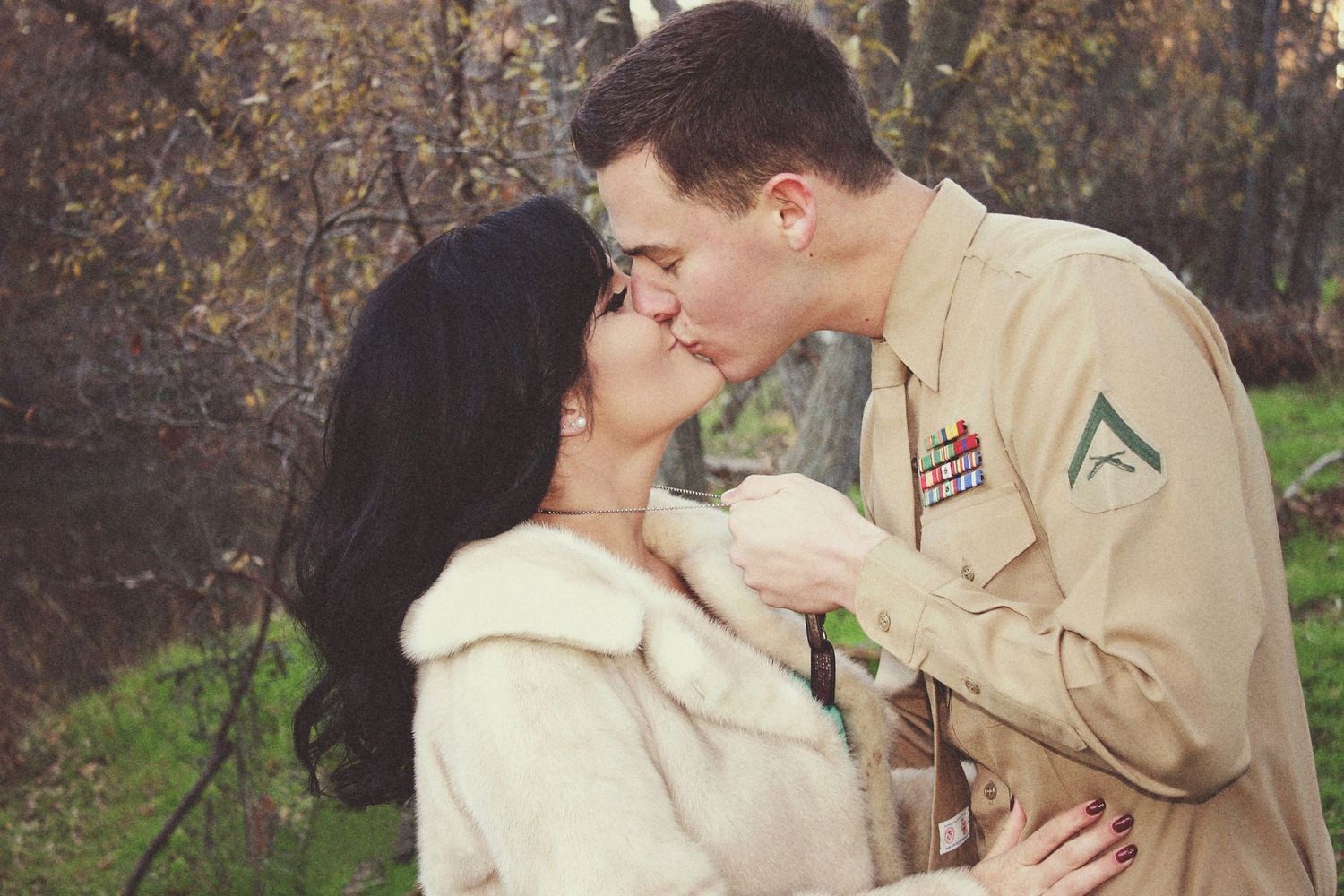 Marine in uniform kissing his dark-haired partner in a white fur coat outdoors among autumn trees.
