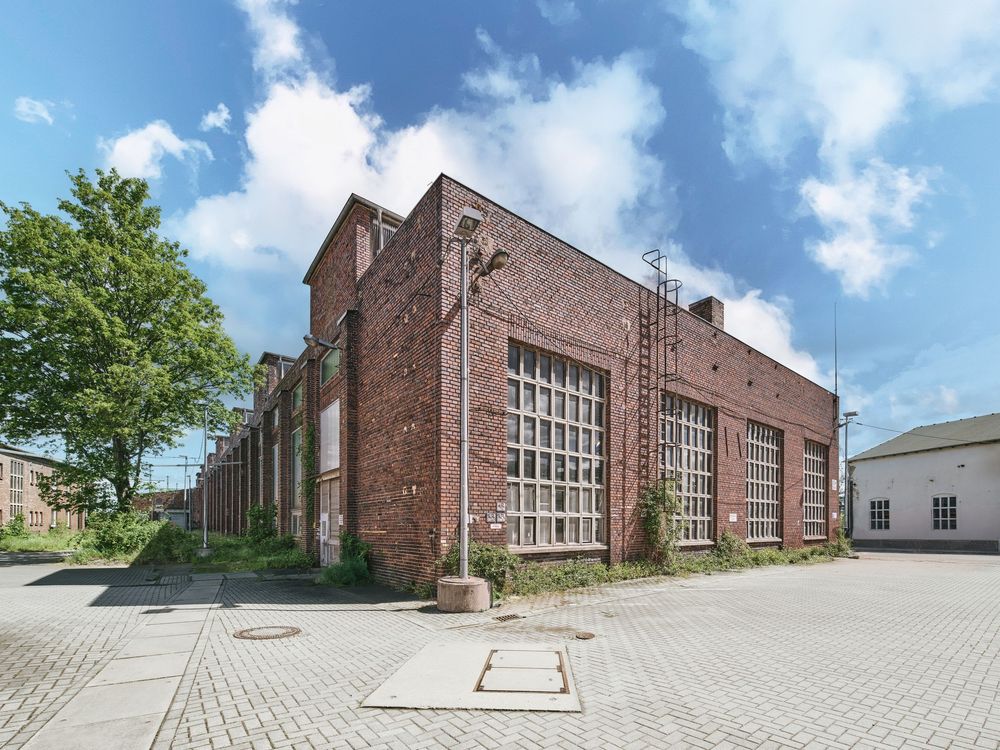 Historic red brick industrial building with large multi-pane windows and a cobblestone courtyard under a bright blue sky with clouds.