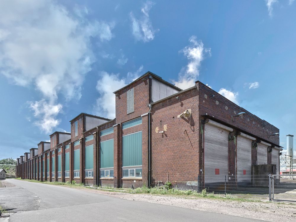 Long red brick industrial building with green-paneled windows and large roll-up doors beside an empty road under a blue sky with clouds.