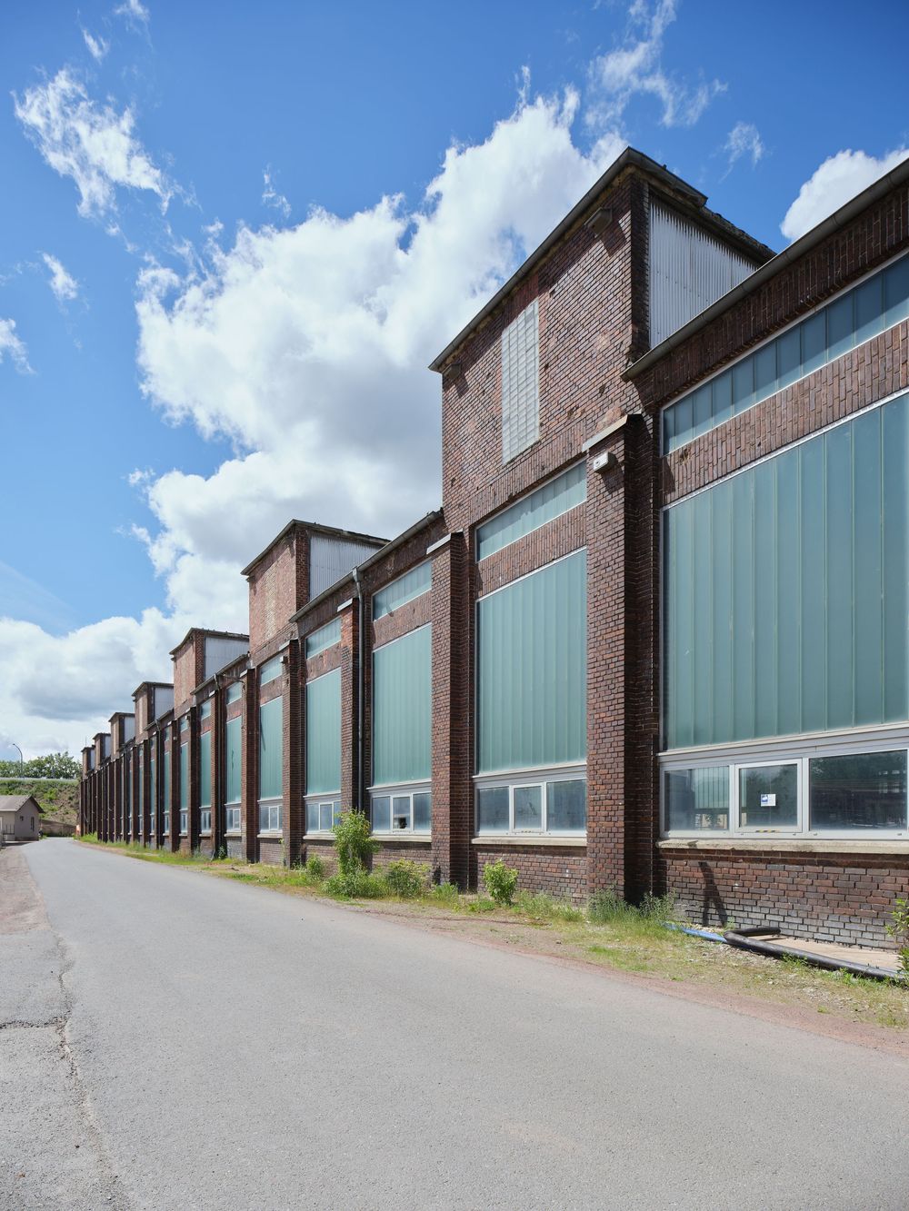 Long red brick industrial building with large green-tinted windows stretching along an empty road under a blue sky with white clouds.