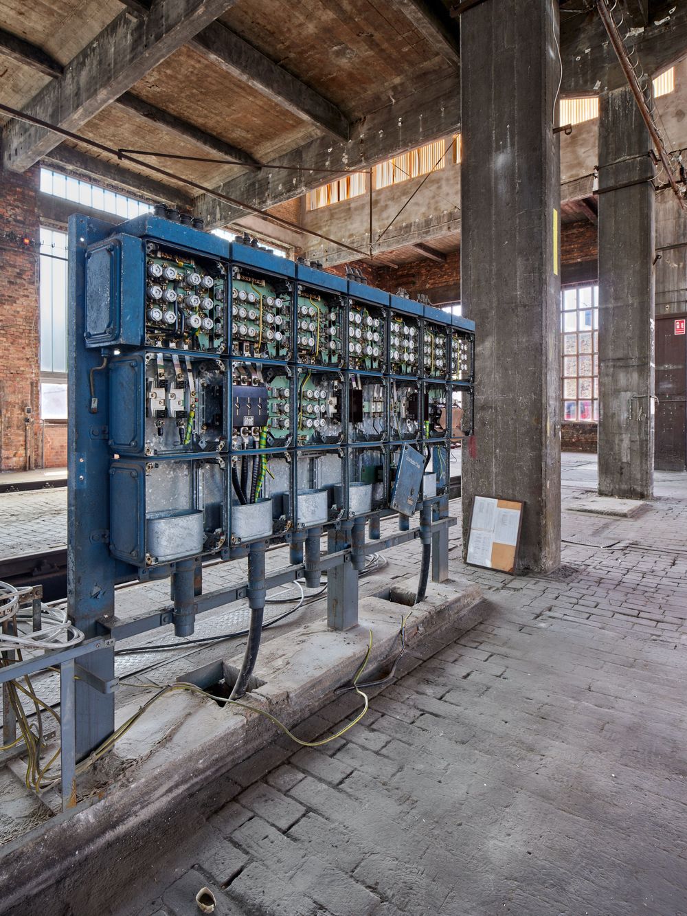 Blue industrial electrical switchboard with wiring and gauges inside an abandoned brick factory hall with tall concrete pillars.