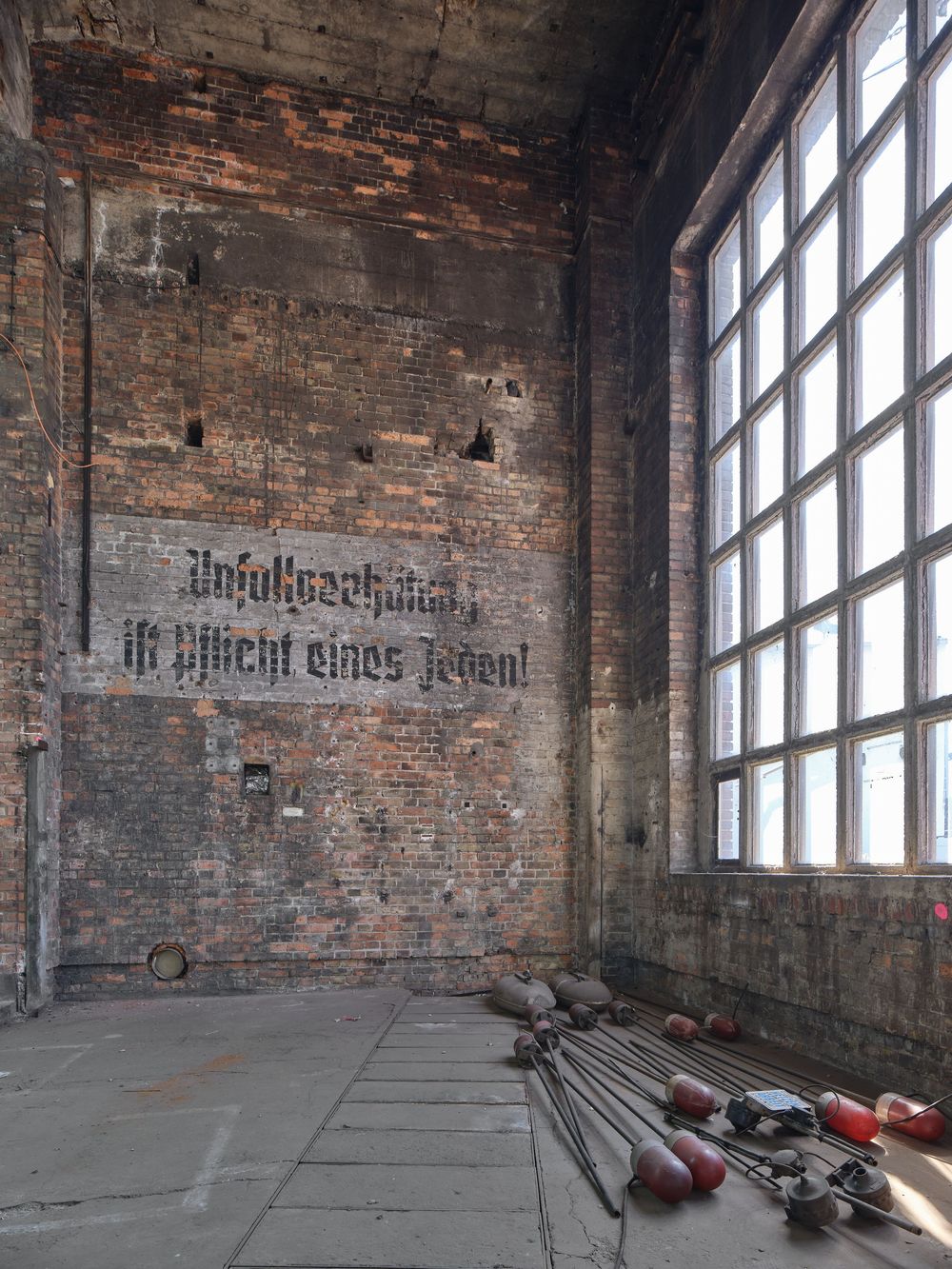 Weathered brick wall with faded German lettering beside a tall factory window, with tools and equipment scattered on the concrete floor.