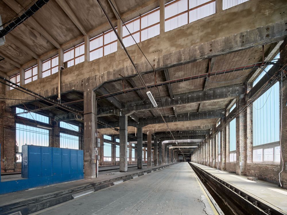 Vast abandoned industrial hall with steel columns, exposed roof trusses, large windows and long tracks stretching into the distance.