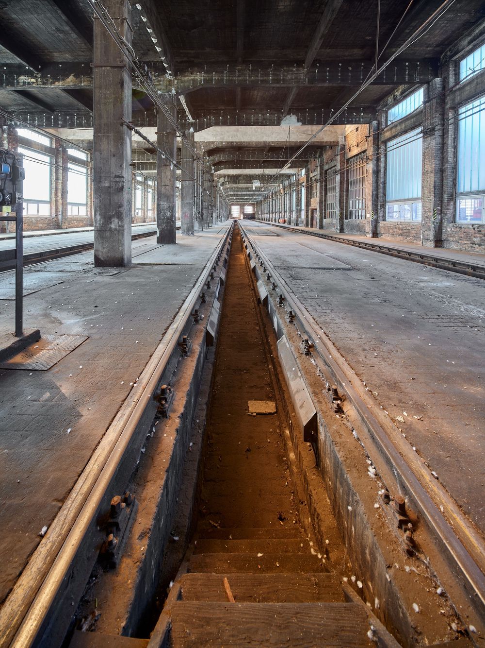 Abandoned industrial hall with a deep maintenance pit running between train tracks, tall windows, and steel support columns.