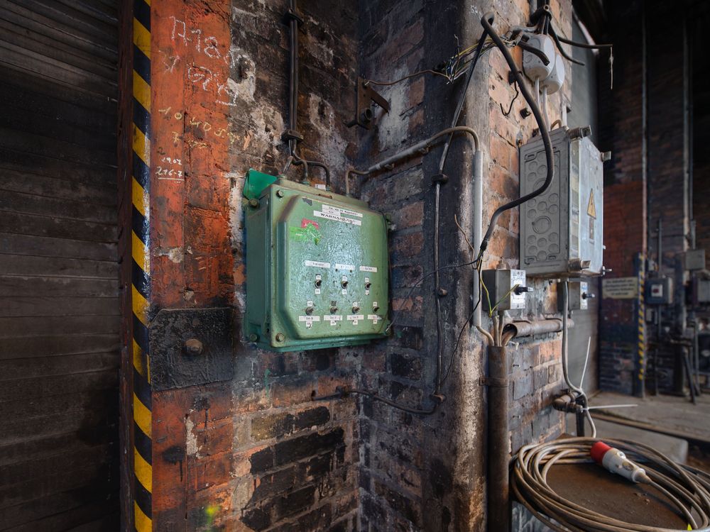 Green electrical control box with switches mounted on a weathered brick wall in a dark industrial building, with coiled cable on the floor.