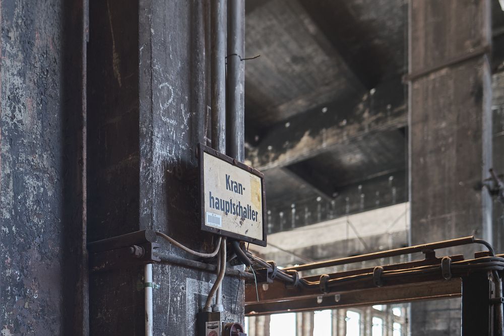 Weathered German warning sign on a dark steel column inside an abandoned industrial hall with high ceilings and rusted beams.