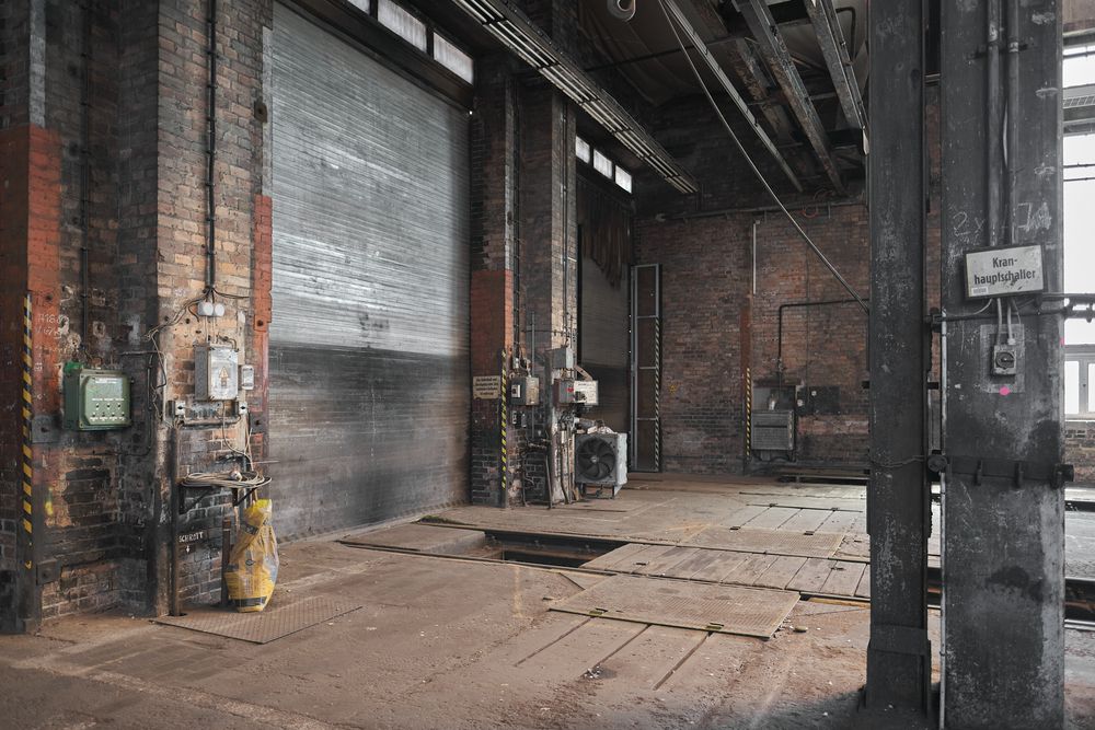 Abandoned industrial warehouse interior with weathered brick walls, a closed roll-up door, rusty pipes, and a worn wooden plank floor.