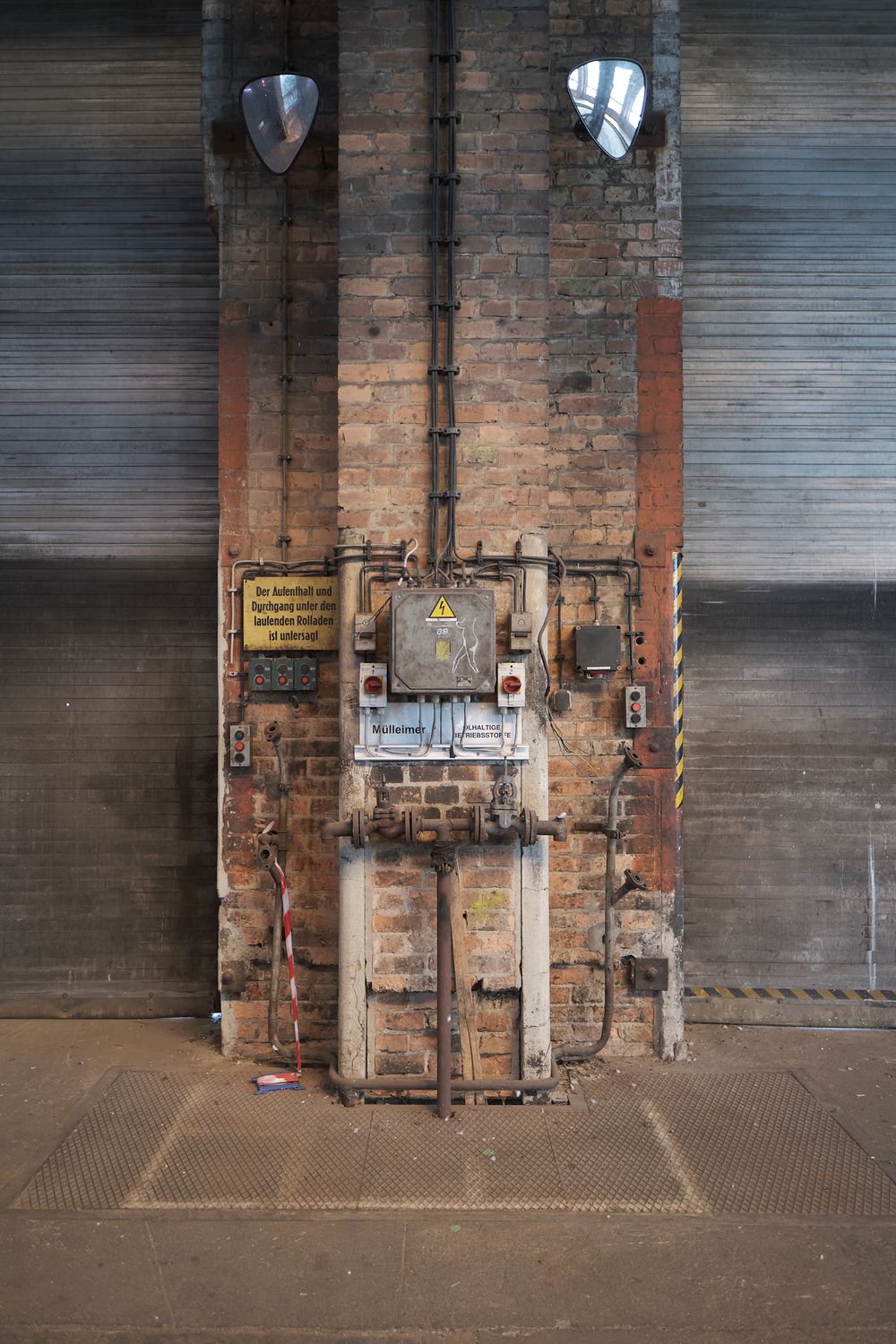Weathered brick pillar in an industrial hall with an electrical control box, pipes, warning signs and corrugated metal walls.