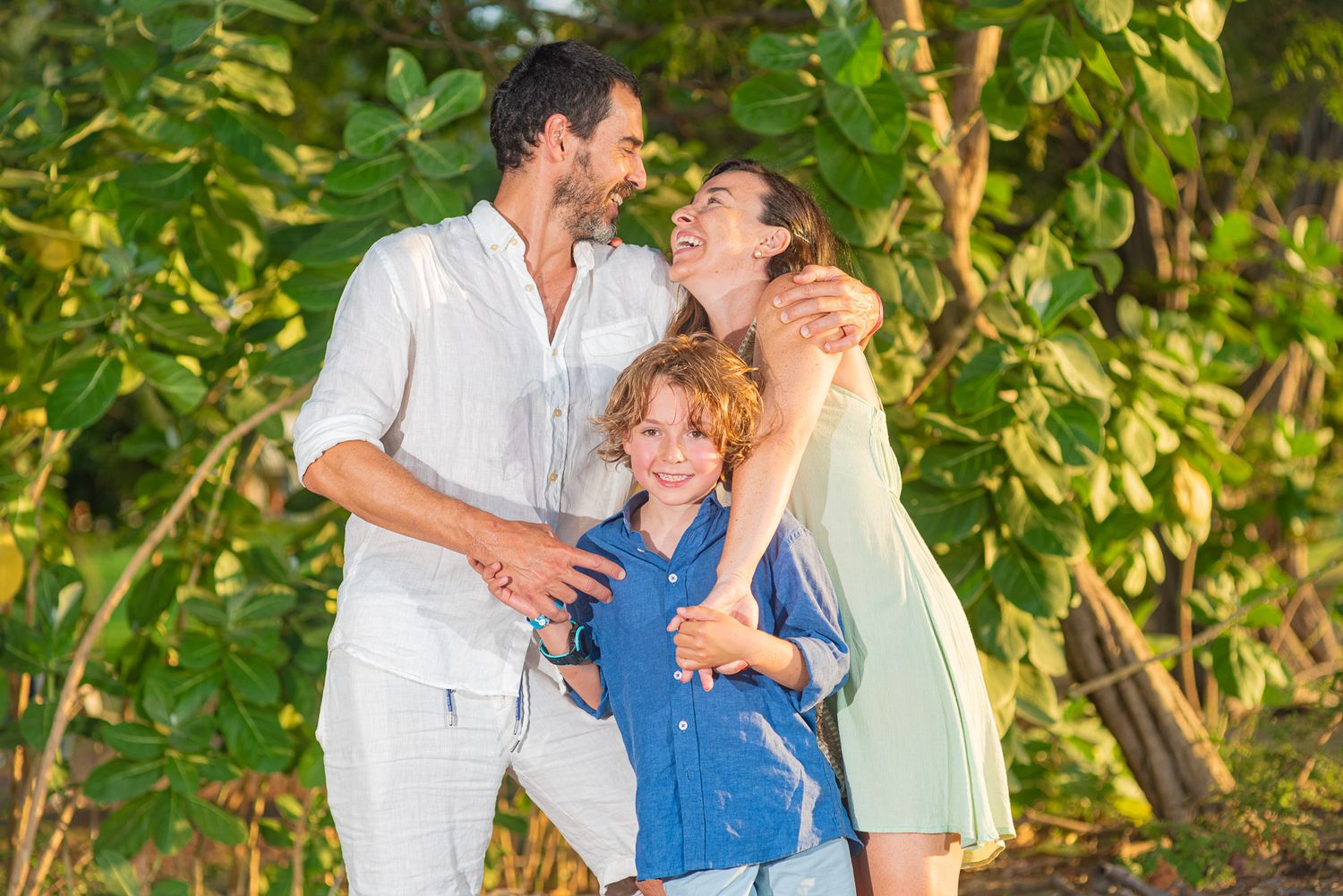 Happy family of three embracing outdoors, a smiling father and mother hugging their young son surrounded by lush green tropical foliage.