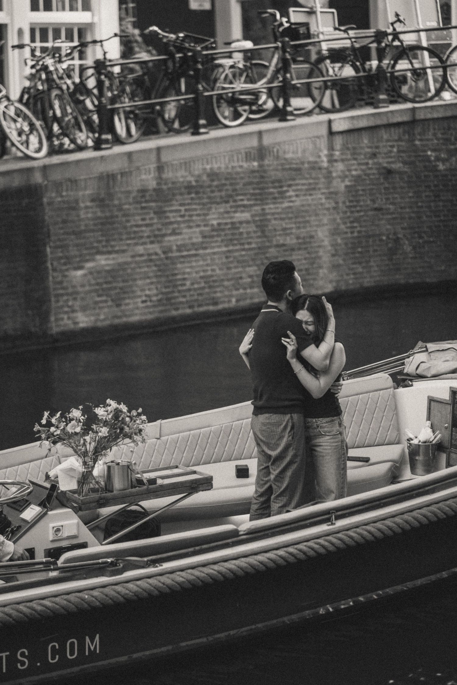 Couple embracing on a boat docked along a canal, with a flower bouquet nearby. Amsterdam secret proposal