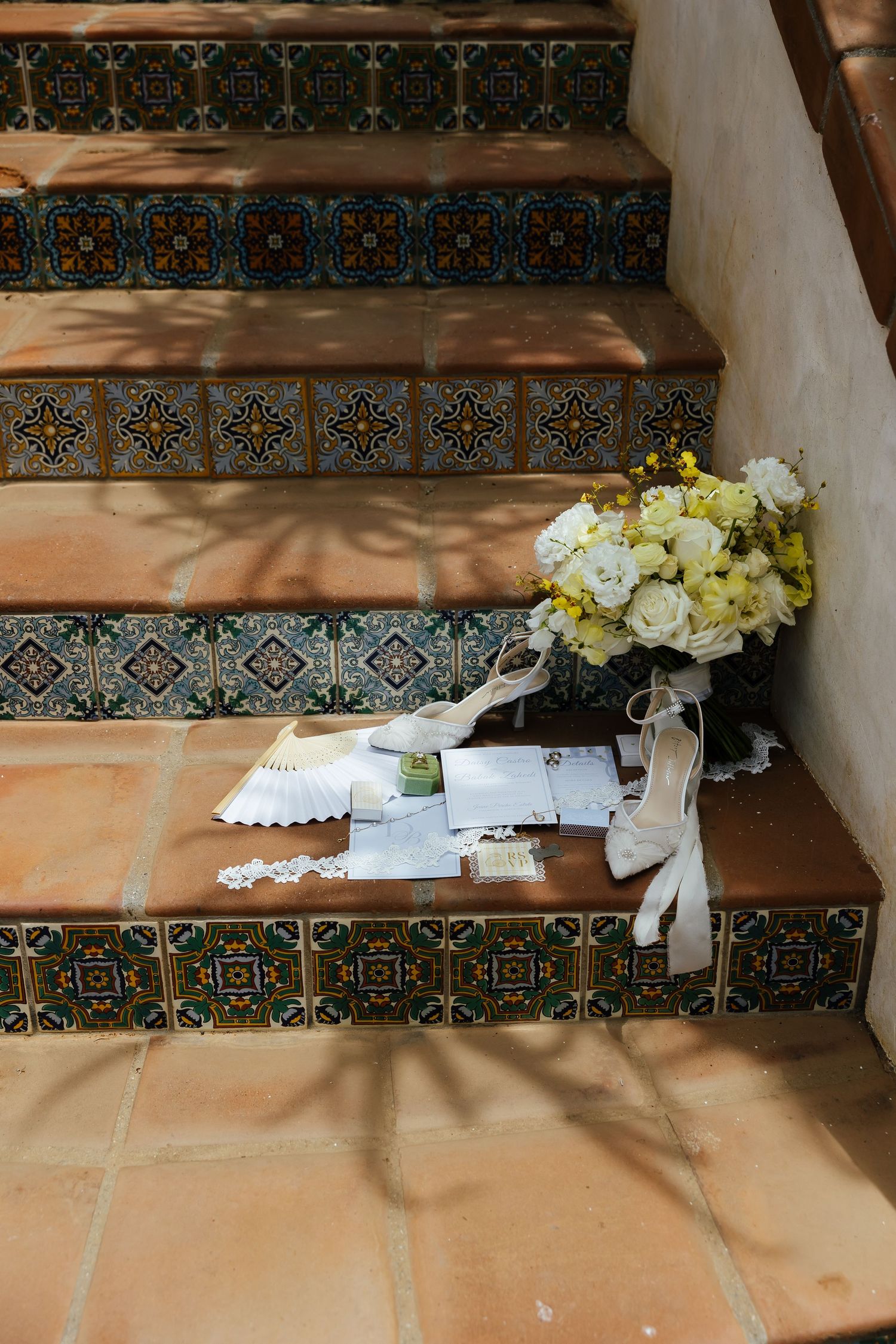 Bridal details including white heels, a lace fan, stationery and a white bouquet arranged on tiled terracotta steps.
