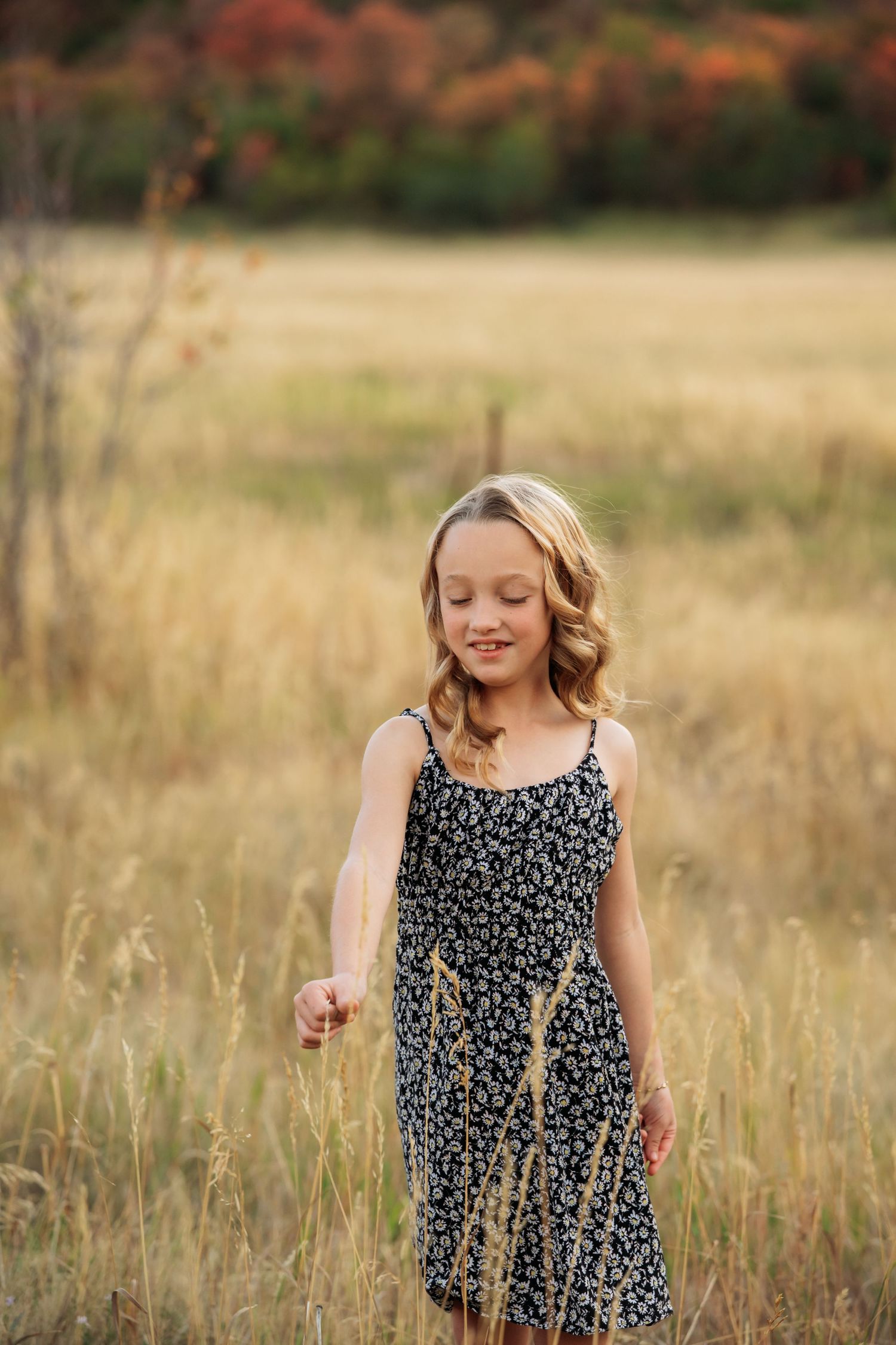Young girl in floral dress crouching in golden field with autumn mountain landscape backdrop.
