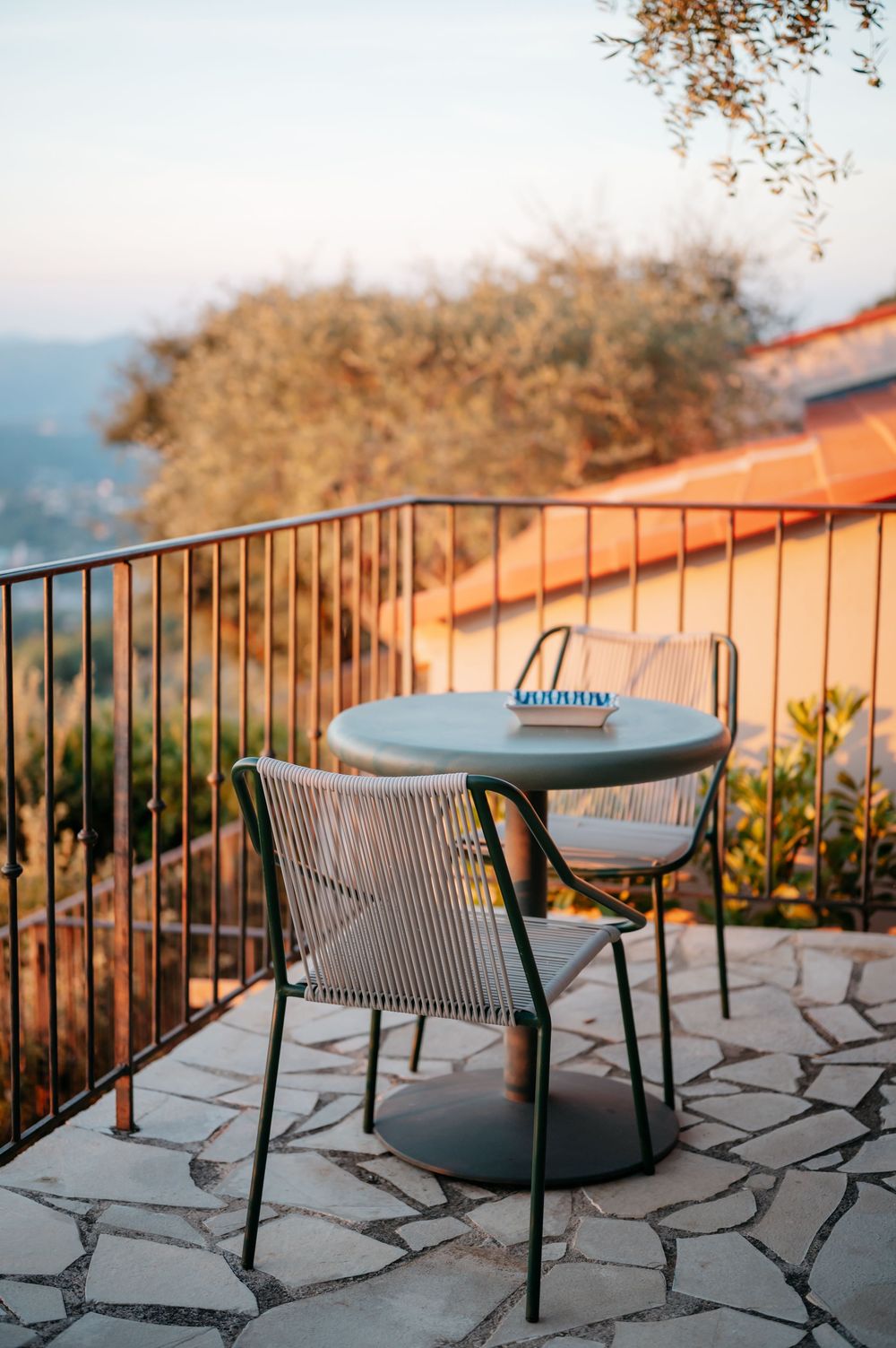 A cozy balcony terrace with a round table and wicker chair on stone flooring, overlooking a scenic hillside at golden hour.