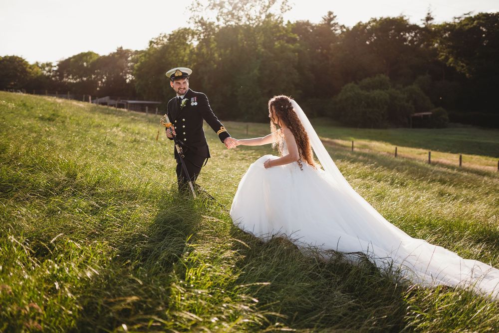 A bride in a white gown and groom in military uniform walk hand in hand through a sunlit green meadow. Tan y Graig Aberystwyth