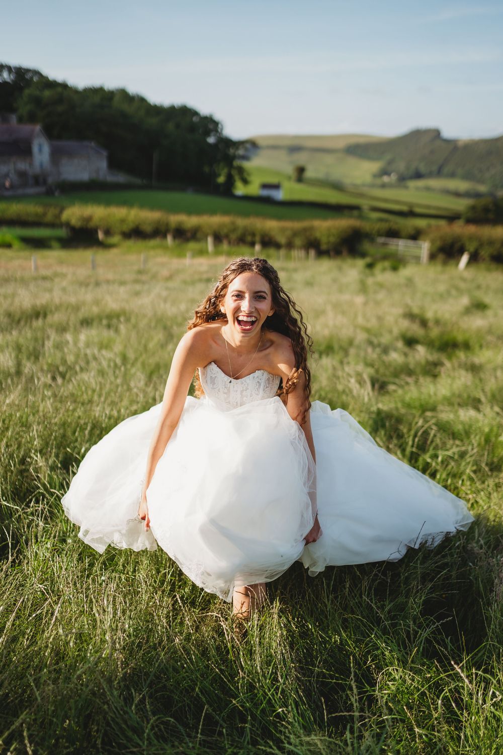 Joyful bride in white strapless ballgown laughing in green countryside field on sunny wedding day.