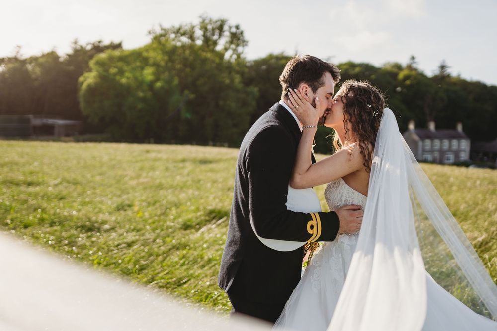 Bride and groom sharing a romantic kiss in a sunlit green field, bride wearing a flowing white gown with veil.