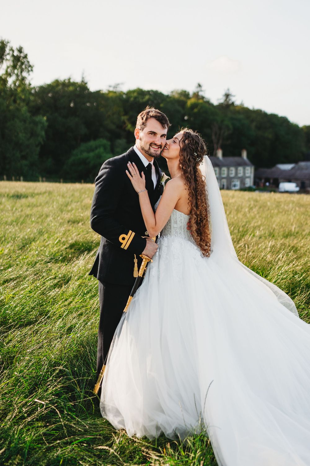 Bride and groom embracing in a sunlit field, groom in military dress uniform, bride in flowing white tulle gown with veil. Tan y Graig Abery