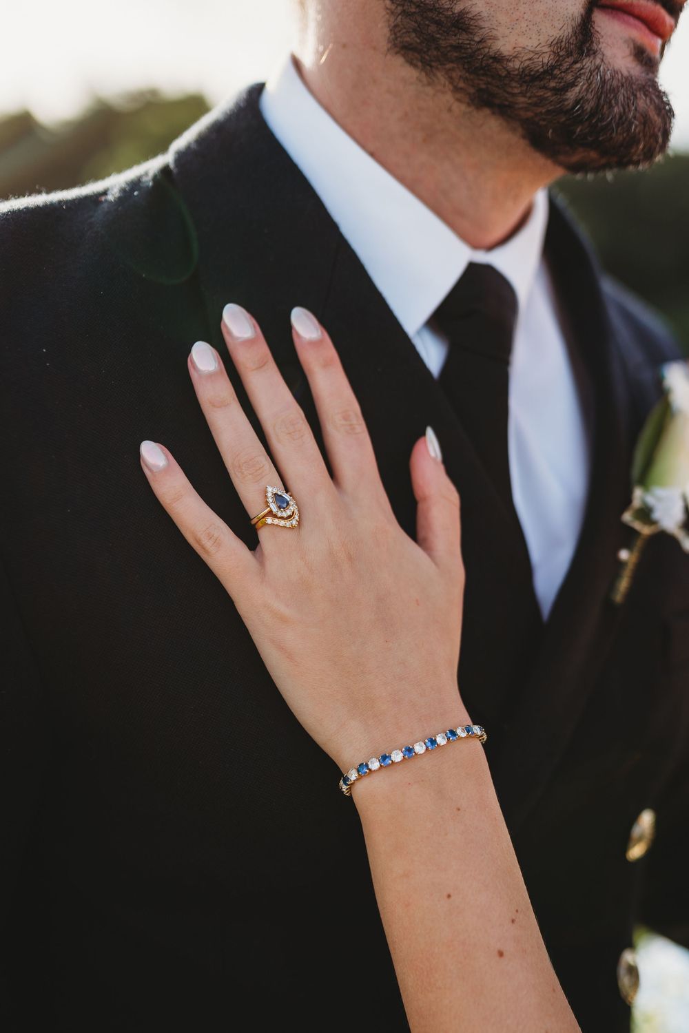 Close-up of a bride's hand displaying a sapphire engagement ring and tennis bracelet against a groom's black suit.