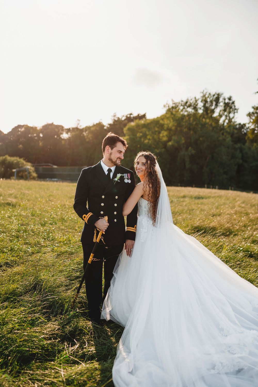 A bride in a white gown and groom in military uniform share a romantic moment in a sunlit green field. Tan y Graig Aberystwyth
