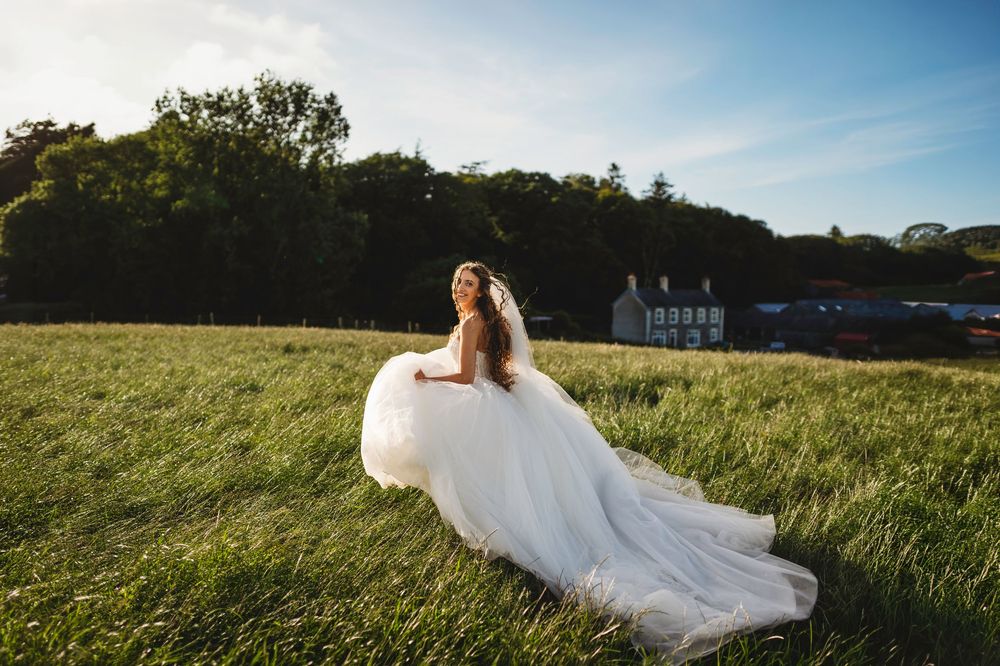 Bride in flowing white tulle gown twirling in sunlit green field with trees and country house backdrop.