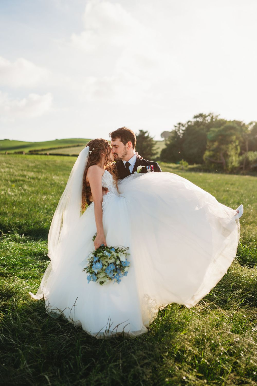 Bride and groom sharing a romantic kiss in a lush green field, bride holding a blue and white bouquet.