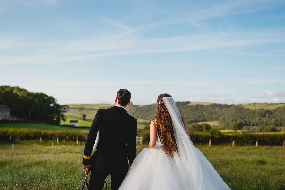 A bride and groom holding hands walking through a lush green countryside field on their wedding day. Tan y Graig Aberystwyth