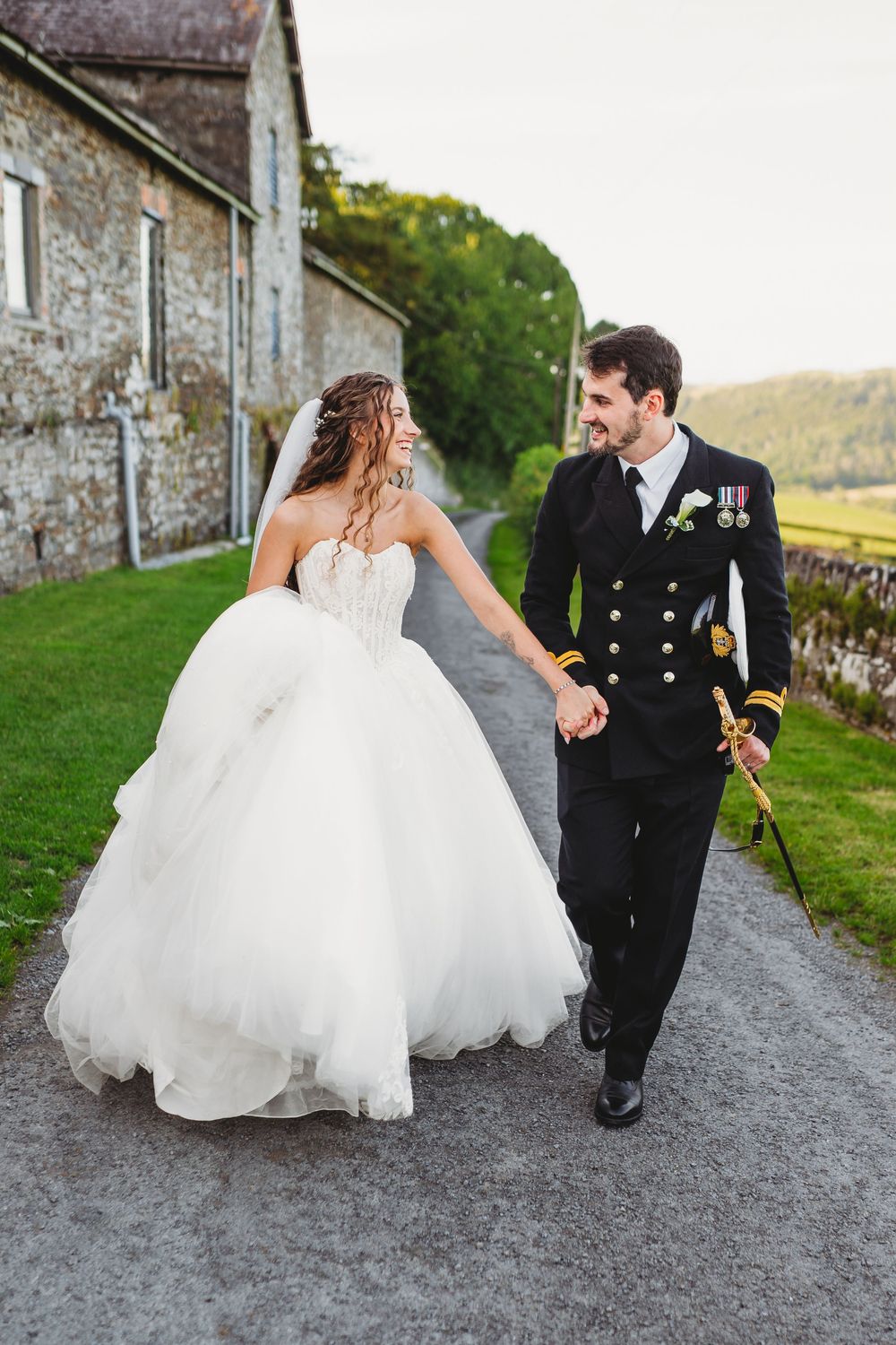 Bride in white ballgown and groom in naval uniform walk hand in hand along a country path beside a stone building. Tan y Graig Aberystwyth