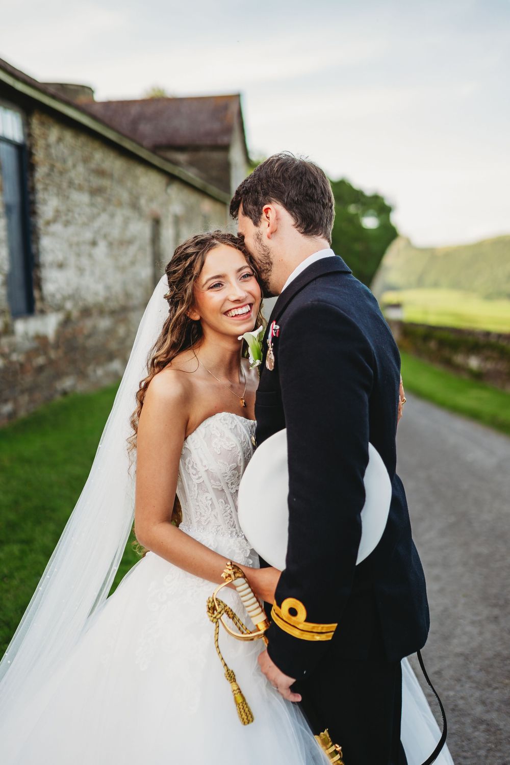 Smiling bride in lace gown and veil embraces groom in military dress uniform outdoors at a countryside wedding venue.