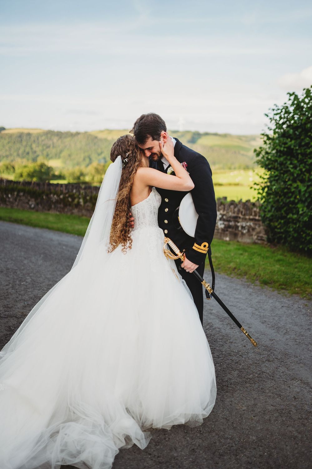 Bride in white gown and groom in military uniform share a romantic kiss on a scenic countryside road.