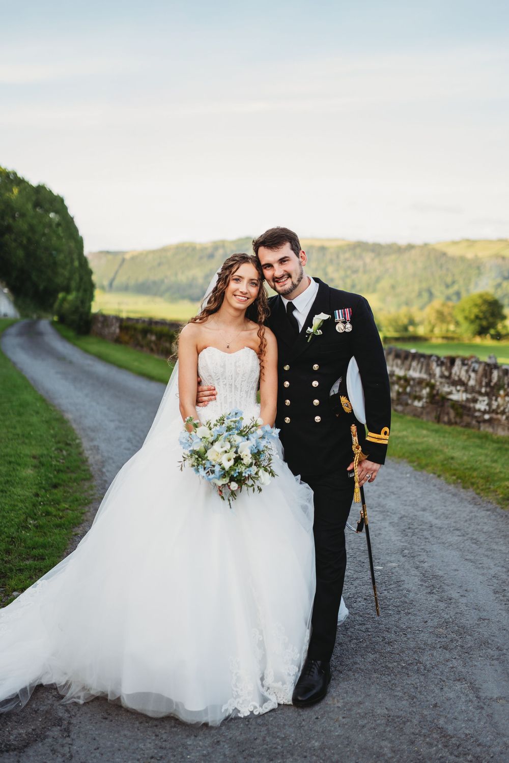 Bride in strapless ball gown holding bouquet stands with groom in naval uniform on a scenic country road.