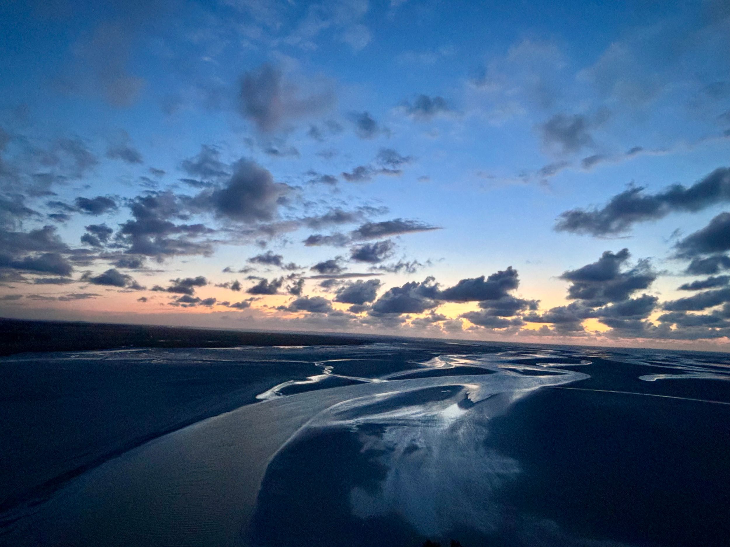 Paysage au crépuscule, avec un ciel bleu traversé de nuages et des teintes orangées à l’horizon. Mont Saint-Michel