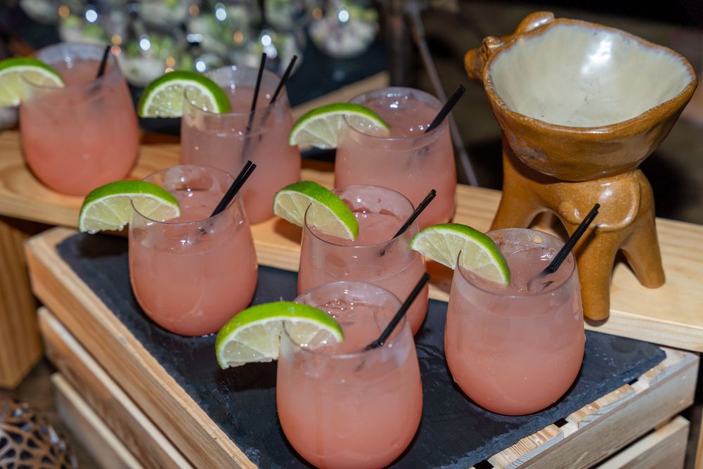 Six pink margaritas garnished with lime wedges and black straws served on a slate board beside a wooden mortar.