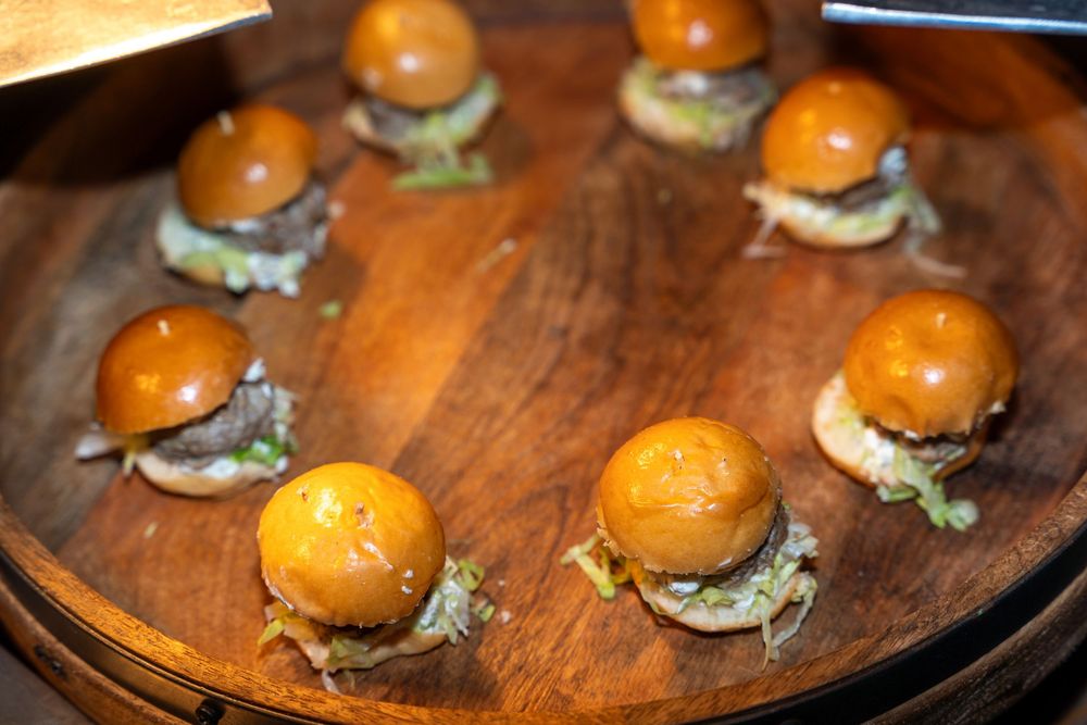 Eight mini sliders with glossy brioche buns arranged on a round wooden serving board.