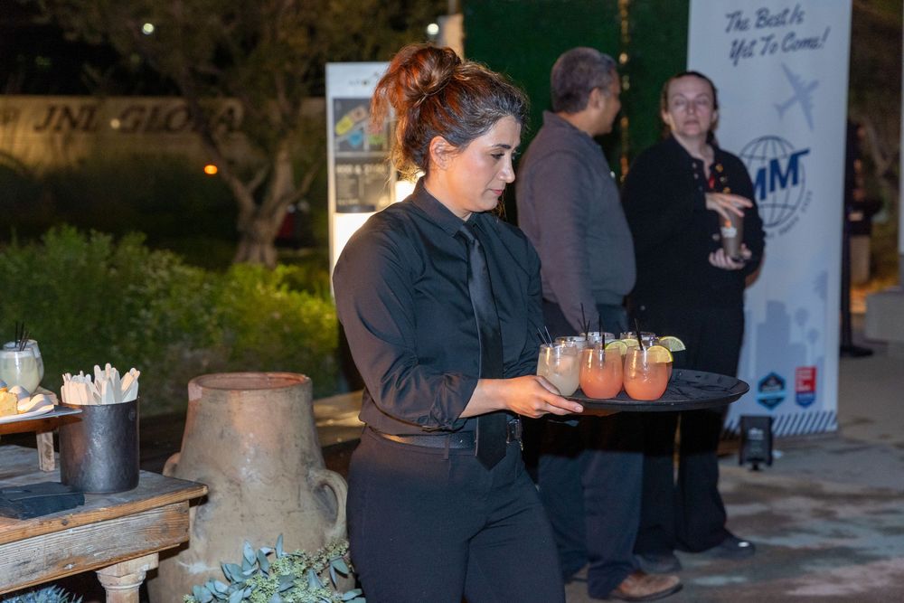 A server in black attire carries cocktails on a tray at an outdoor catered evening event.