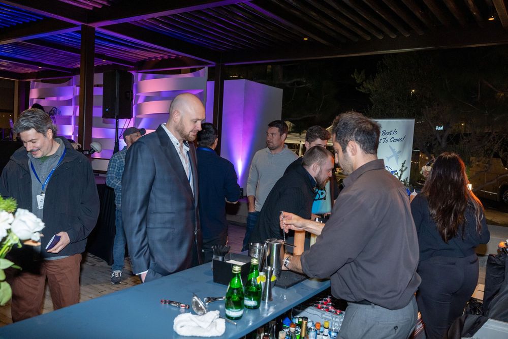 Guests mingle at an outdoor evening networking event with a bar setup, ambient purple lighting, and lush greenery.